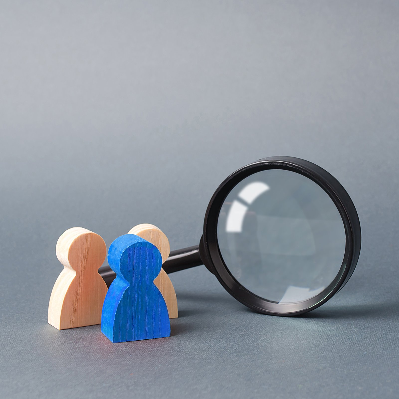 Three wooden human figure stands near a magnifying glass on a gray background.
